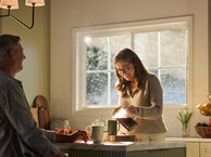 A warmly lit kitchen featuring a large Restorations Series window that frames a soft, sunlit outdoor view. A woman wearing glasses smiles as she pours coffee from a glass carafe into a mug on a marble kitchen island. A man sits across from her, partially visible, with a mug and fresh strawberries nearby. The space includes light cabinetry, a fruit basket on the counter, and gentle morning sunlight streaming through the multi-pane window.