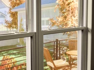 View through a double-pane window onto a small outdoor deck with a wooden table and chairs, framed by autumn foliage and neighboring houses in soft daylight.