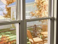 View through a double-pane window onto a small outdoor deck with a wooden table and chairs, framed by autumn foliage and neighboring houses in soft daylight.