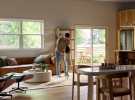 A bright, modern living and dining area featuring large Vanguard Series windows that fill the space with natural light and views of lush greenery outside. A man stands near the center of the room playfully holding his medium-sized dog upright on its hind legs. The room includes a brown leather sofa with accent pillows, a round white coffee table with plants and books, a wooden dining table with chairs, and a sliding glass door leading to a small outdoor patio with additional seating.