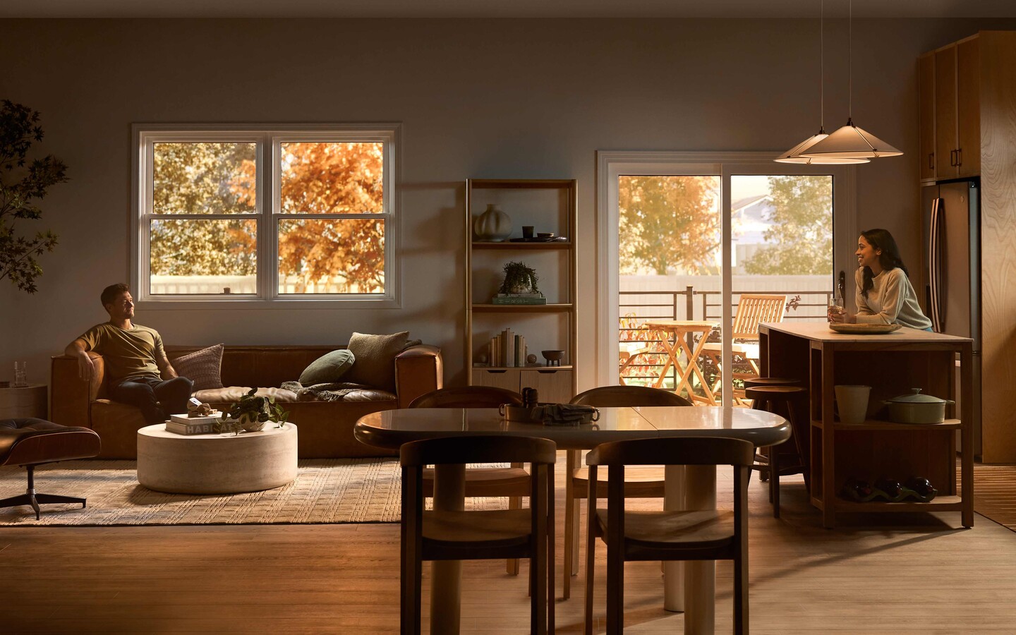 A wide view of an open-plan living room and kitchen where a man relaxes on a sofa and a woman sits at a kitchen island, with warm wood cabinetry, modern furniture, pendant lights, and large windows showing autumn trees and an outdoor deck.