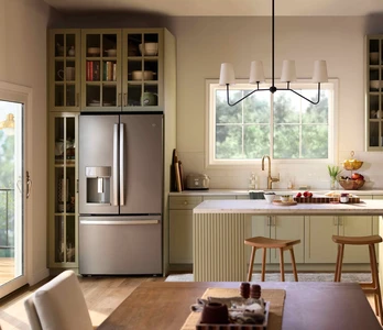 A bright, modern kitchen with sage-green cabinetry, a central island with stools, stainless steel refrigerator, and open shelving, illuminated by natural light from a Restorations Series Horizontal sliding window and a glass door leading to an outdoor deck.
